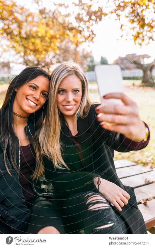 Two smiling young women on park bench taking a selfie Selfie Selfies woman females parks benches smile female friends mobile phone mobiles mobile phones