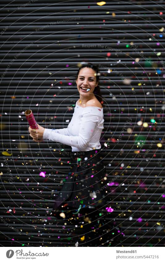 Happy young woman standing in front of black roller shutter throwing confetti females women Adults grown-ups grownups adult people persons human being humans
