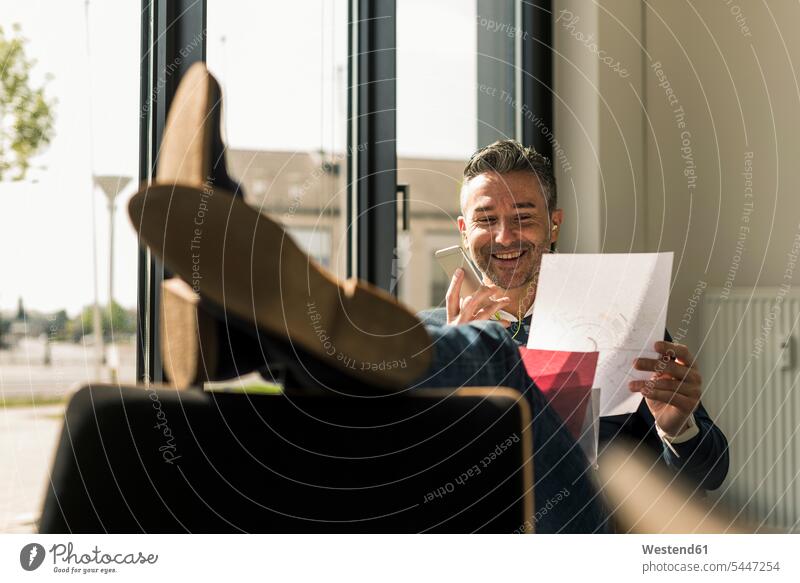 Smiling businessman sitting with feet up in his office using smartphone and earphones on the phone call telephoning On The Telephone calling Businessman