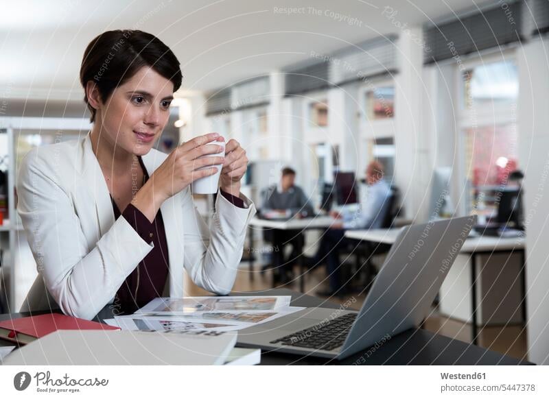 Woman using laptop in office with colleagues in background Laptop Computers laptops notebook businesswoman businesswomen business woman business women females