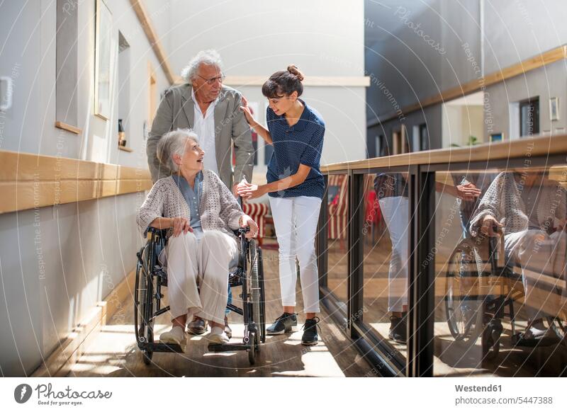 Gereatric nurse helping senior man and woman to walk down corridor geriatric nurse wheelchair wheel chair helpfulness together talking speaking assistance
