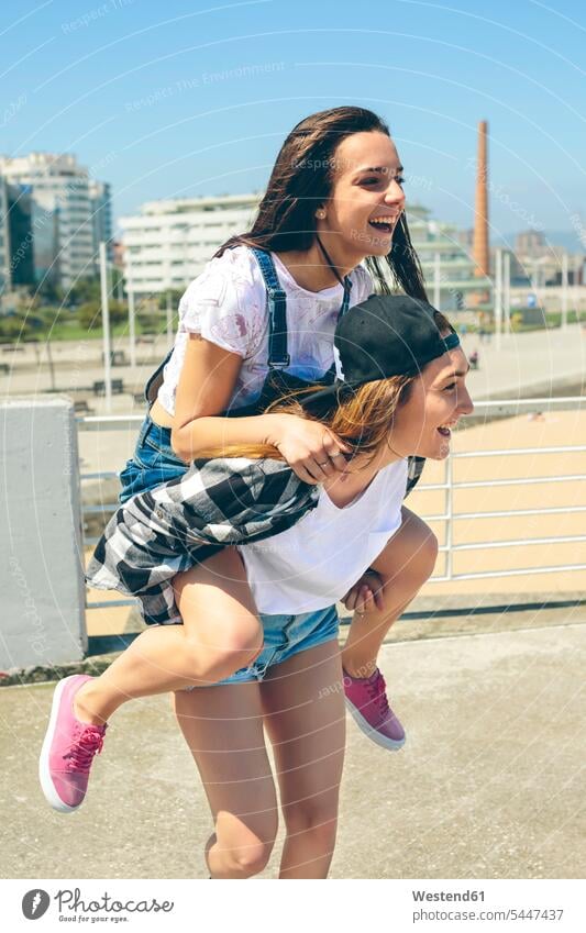 Young woman giving her friend a piggyback ride on roof terrace piggy-back pickaback Piggybacking Piggy Back female friends mate friendship laughing Laughter