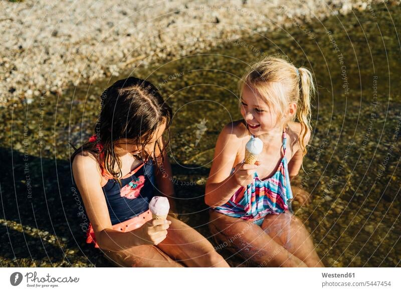 Two girls sitting in water at lakeshore eating icecream females female friends child children kid kids people persons human being humans human beings mate