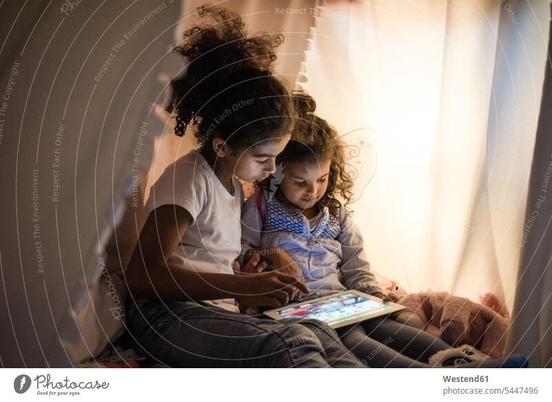 Two sisters sitting in dark children's room, looking at digital tablet playing Kids Room nursery child's room reading Seated rooms domestic room domestic rooms