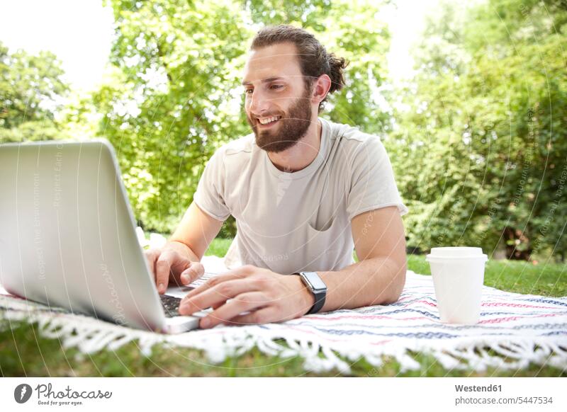 Smiling man with coffee to go lying on blanket in a park using laptop Laptop Computers laptops notebook men males computer computers Adults grown-ups grownups