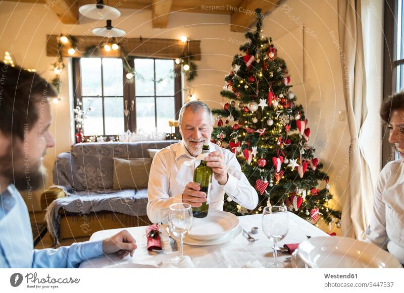Smiling senior man with family holding bottle of wine at Christmas dinner table X-Mas yule Xmas X mas Wine families smiling smile celebrating celebrate partying