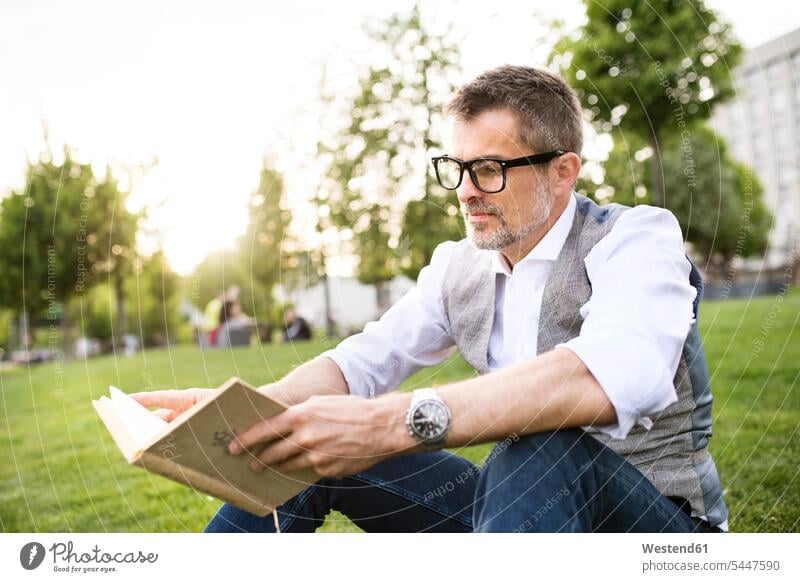 Confident mature businessman in the city park sitting on grass reading book meadow meadows books relaxed relaxation men males relaxing Adults grown-ups grownups