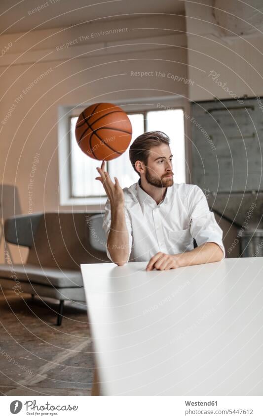 Portrait of pensive young businessman sitting at table in a loft balancing basketball on his finger Businessman Business man Businessmen Business men