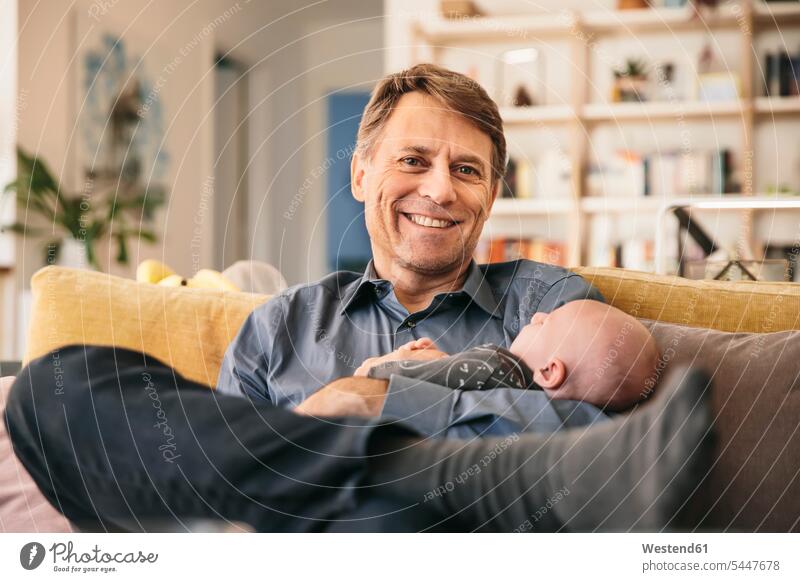 Smiling father and his newborn baby sitting on couch at home settee sofa sofas couches settees pa fathers daddy dads papa babies infants smiling smile parents