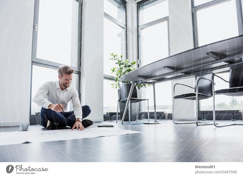 Smiling businessman sitting on the floor of his office looking at construction plan Businessman Business man Businessmen Business men business people