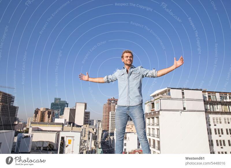 Young man standing on rooftop terrace with arms outstretched men males happiness happy blond blond hair blonde hair roof terrace deck carefree smiling smile