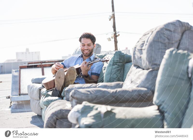 Young man on rooftop sitting on sofa and playing guitar musician musicians cheerful gaiety Joyous glad Cheerfulness exhilaration merry gay couch settee sofas