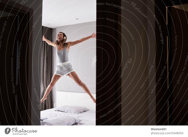 Excited woman wearing headphones jumping in bed Leaping headset beds females women excitement enthusiastic enthusiasm excited Ardour Ardor jumps Adults