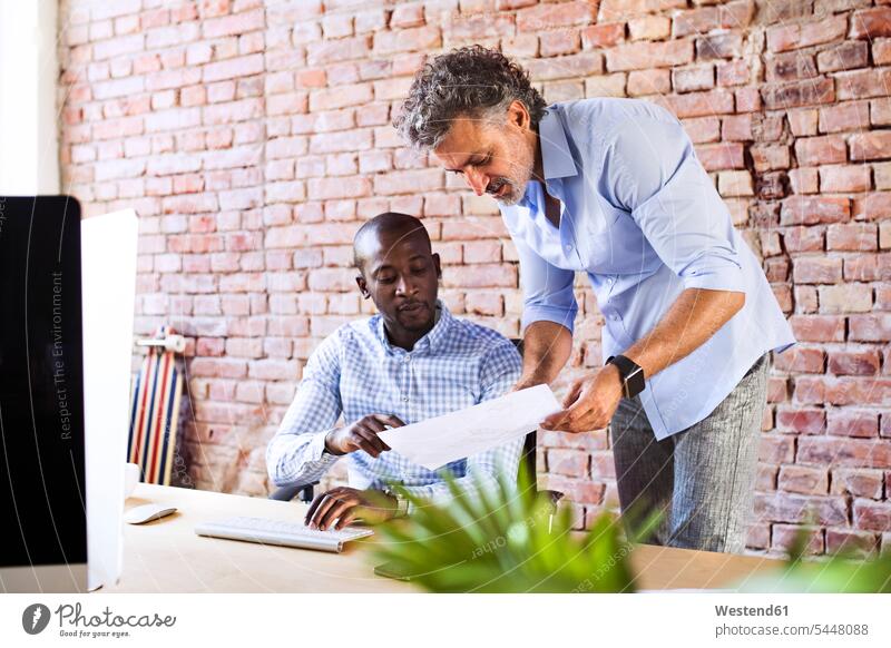 Two colleagues looking at paper at desk in office offices office room office rooms working At Work together desks workplace work place place of work Table