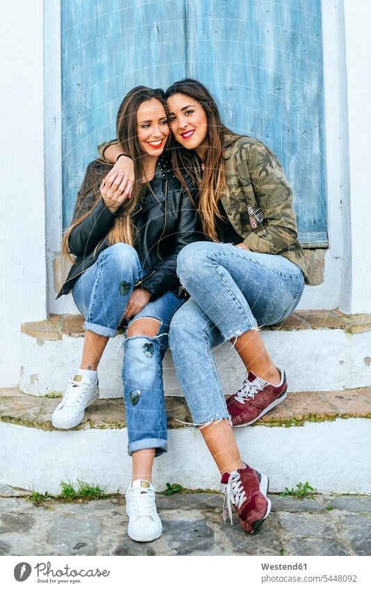 Two smiling young women sitting on stoop hugging female friends embracing embrace Embracement Seated smile mate friendship woman females Adults grown-ups