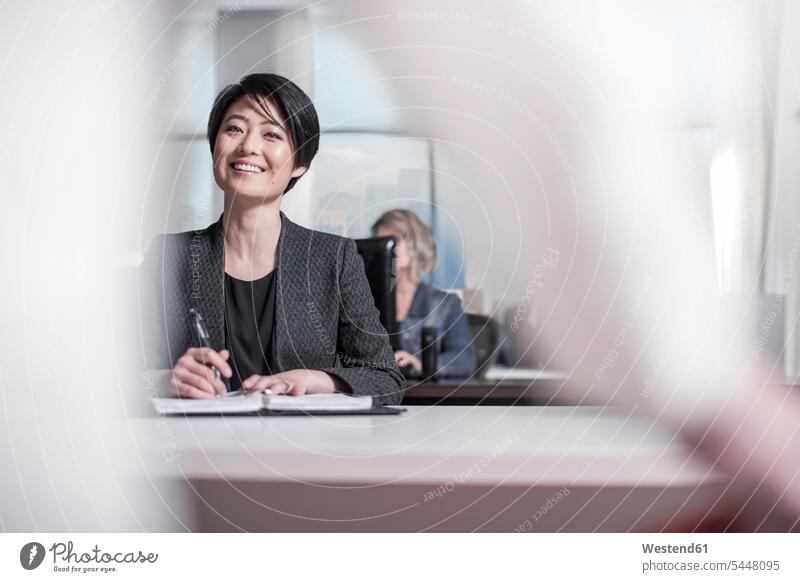 Smiling woman sitting at desk in city office notebook notebooks smiling smile colleagues talking speaking offices office room office rooms workplace work place