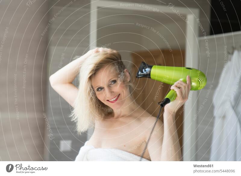 Portrait of smiling blond woman using hair dryer in the bathroom portrait portraits Bath females women blow-drying blow drying hair Adults grown-ups grownups