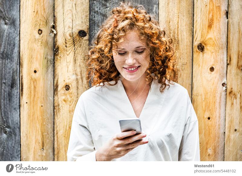 Portrait of smiling young woman looking at cell phone in front of wooden wall females women Smartphone iPhone Smartphones portrait portraits Adults grown-ups