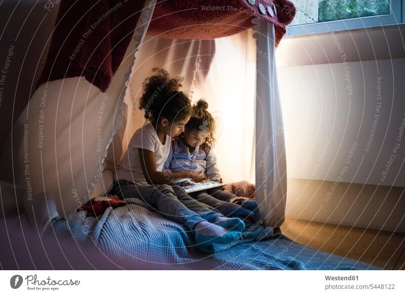 Two sisters sitting in dark children's room, looking at digital tablet Seated playing reading Kids Room nursery child's room siblings brother and sister