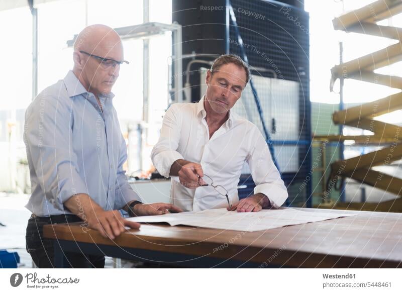 Two businessmen looking at plan on table in factory factories plans Businessman Business man Businessmen Business men talking speaking business people