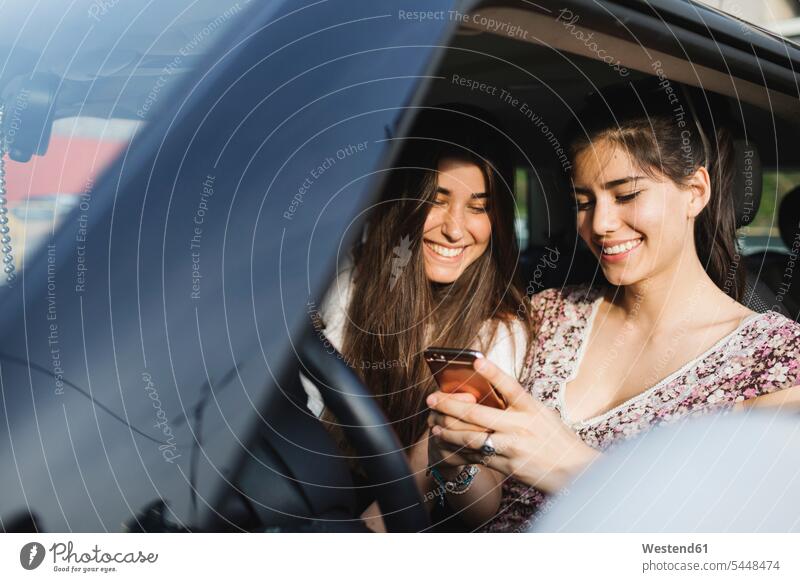 Two happy young women looking at cell phone in a car smiling smile female friends automobile Auto cars motorcars Automobiles mobile phone mobile phones