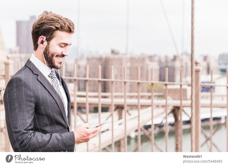 USA, New York City, smiling businessman with cell phone and earbuds on Brooklyn Bridge bridge bridges mobile phone mobiles mobile phones Cellphone cell phones