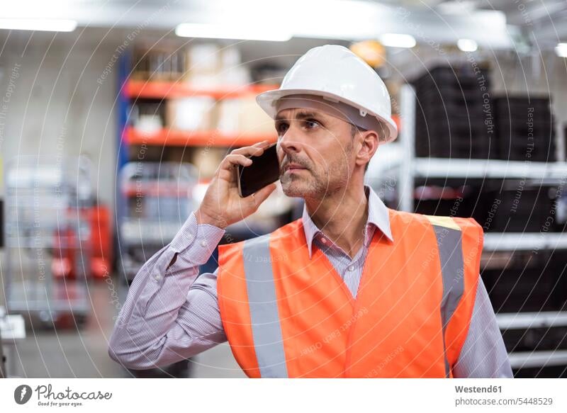 Man in factory hall wearing safety vest and hard hat talking on cell phone man men males on the phone call telephoning On The Telephone calling mobile phone