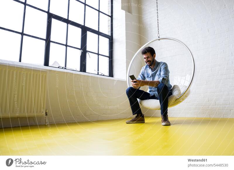 Young man sitting on swing in his loft looking at cell phone Smartphone iPhone Smartphones men males mobile phone mobiles mobile phones Cellphone cell phones