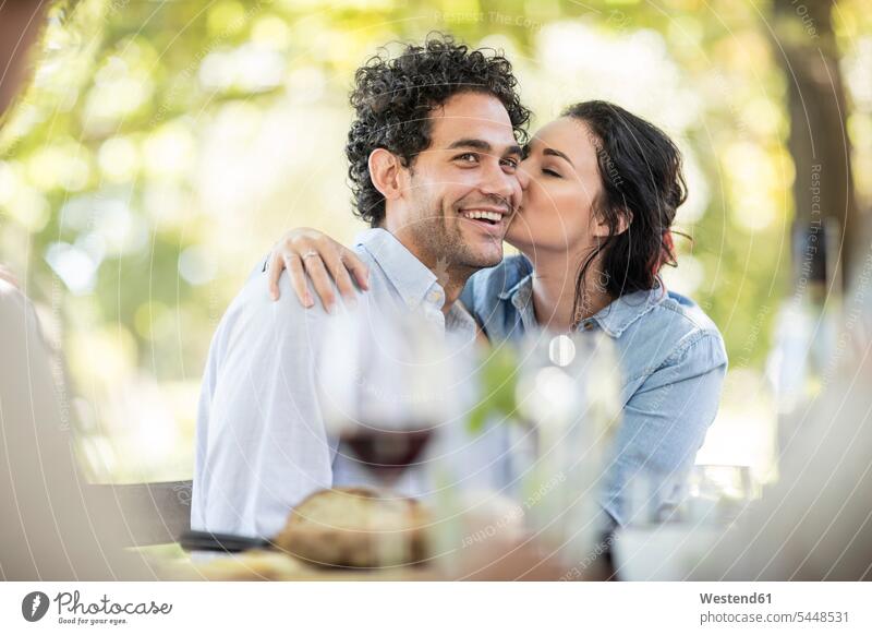 Young woman kissing a happy young man at outdoor table Wine smiling smile kisses Red Wine Red Wines couple twosomes partnership couples celebrating celebrate