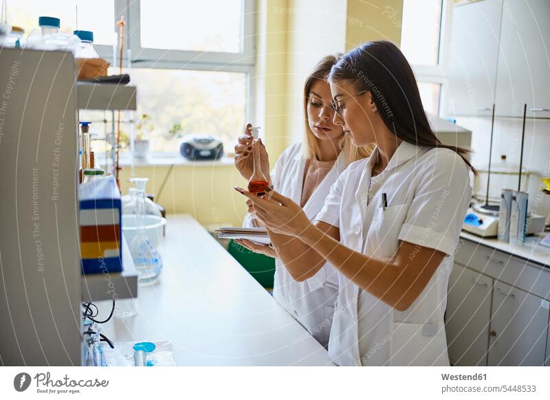 Two young women looking at liquid in flask in laboratory eyeing woman females recipient Chemical Flasks together liquids view seeing viewing Adults grown-ups