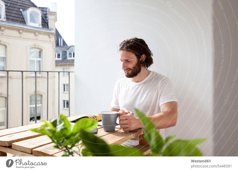 Smiling man sitting with coffee mug on balcony reading magazine journal magazines journals men males balconies Adults grown-ups grownups adult people persons