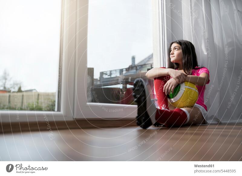 Girl in soccer outfit sitting on floor in living room looking outside window Seated girl females girls soccer ball soccer balls footballs child children kid