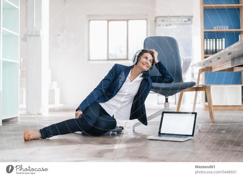 Smiling businesswoman sitting on the floor in a loft listening music with headphones businesswomen business woman business women laptop Laptop Computers laptops
