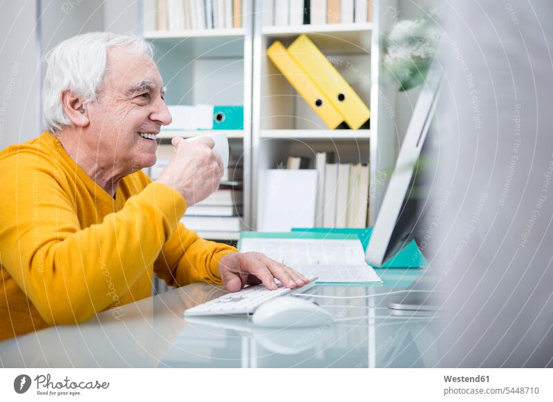Senior man working at computer with a cup of coffee sitting Seated At Work drinking Coffee computers men males senior men senior man elder man elder men
