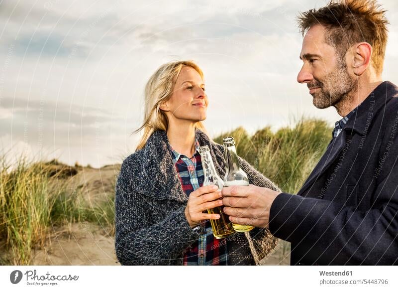 Smiling couple in dunes clinking beer bottles beach beaches twosomes partnership couples sand dune sand dunes smiling smile Beer Beers Ale people persons