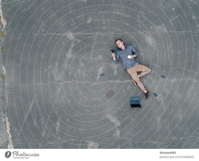 Young businessman with coffee to go lying on tarmac using mini tablet, quadcopter view Businessman Business man Businessmen Business men use asphalt Blacktop
