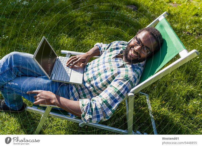 Smiling man with laptop sitting on deck chair on a meadow looking up to camera men males Laptop Computers laptops notebook deckchair deckchairs deck chairs