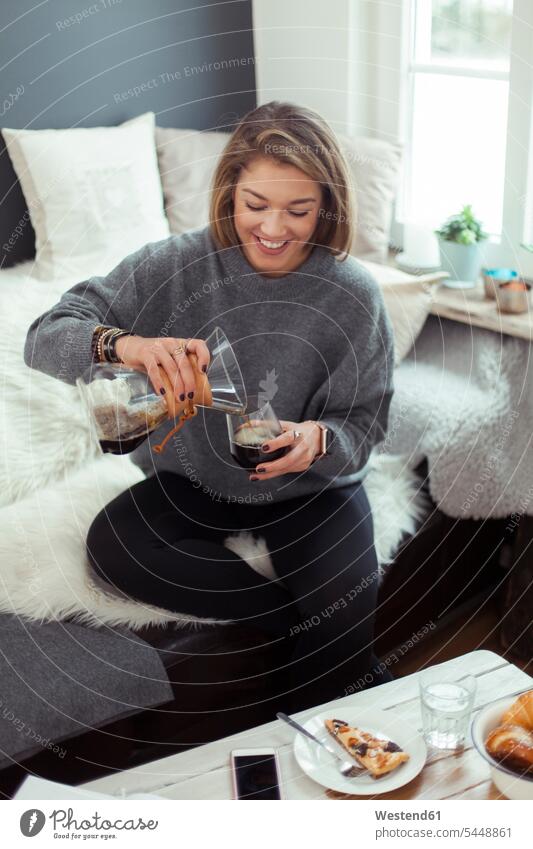 Smiling blond woman sitting on the couch pouring coffee into a glass Coffee females women Drink beverages Drinks Beverage food and drink Nutrition Alimentation