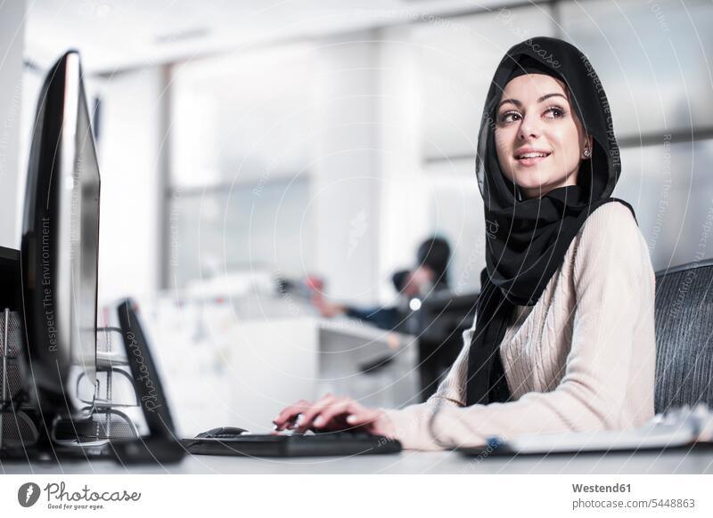 Young woman wearing hijab working on desk in office smiling smile females women portrait portraits offices office room office rooms At Work headscarf head scarf
