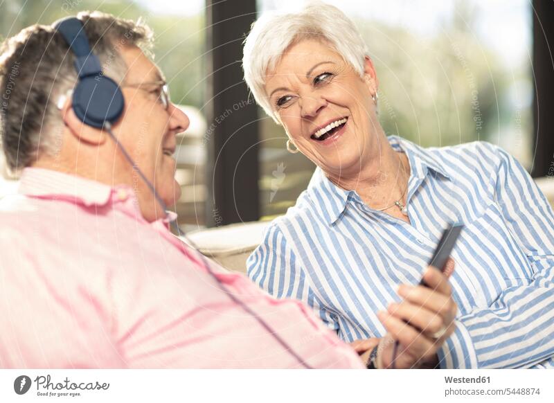 Happy senior couple with cell phone and headphones on couch at home headset happiness happy twosomes partnership couples hearing settee sofa sofas couches