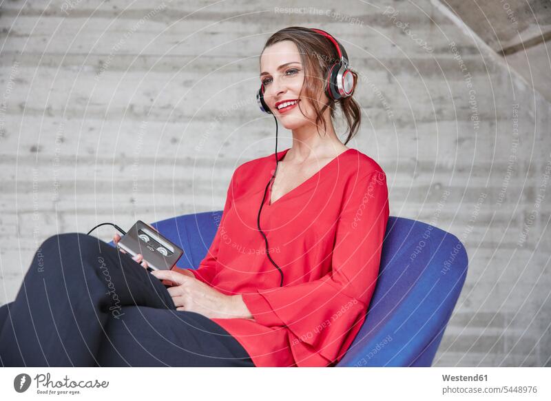 Smiling woman sitting on chair listening to music from walkman Seated smiling smile headphones headset females women hearing Adults grown-ups grownups adult