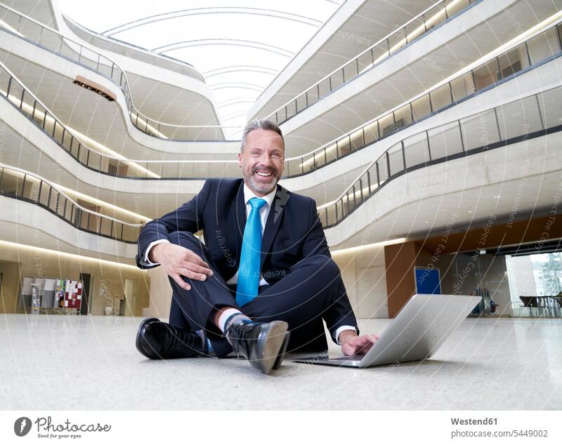 Happy businesssman sitting on floor in modern office building using laptop Seated smiling smile Laptop Computers laptops notebook Businessman Business man