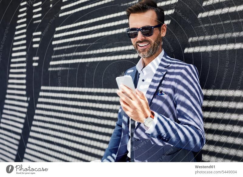 Businessman wearing sunglasses using phone near concrete wall smiling smile Business man Businessmen Business men mobile phone mobiles mobile phones Cellphone