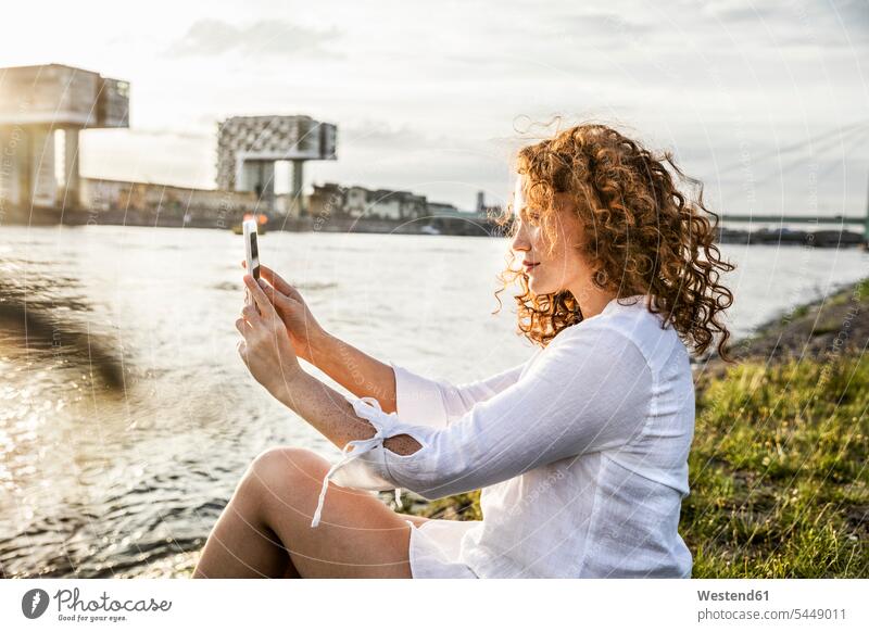 Germany, Cologne, young woman sitting at riverside in the evening taking selfie with cell phone females women Selfie Selfies Adults grown-ups grownups adult