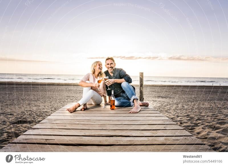 Happy couple with wine glasses sitting on boardwalk on the beach at sunset Wine twosomes partnership couples Seated beaches relaxed relaxation Alcohol