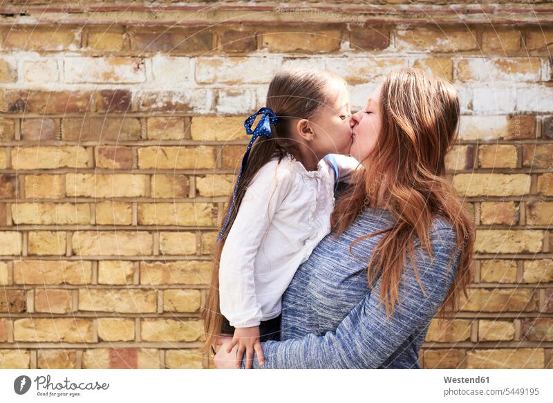 Mother kissing her little daughter in front of brick wall daughters kisses mother mommy mothers ma mummy mama child children family families people persons