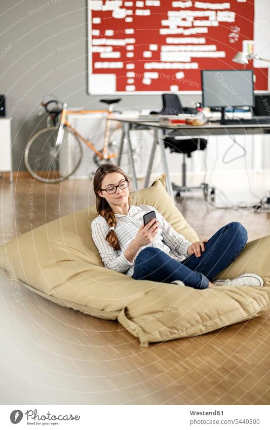 Young woman with cell phone sitting in bean bag in office Seated offices office room office rooms mobile phone mobiles mobile phones Cellphone cell phones