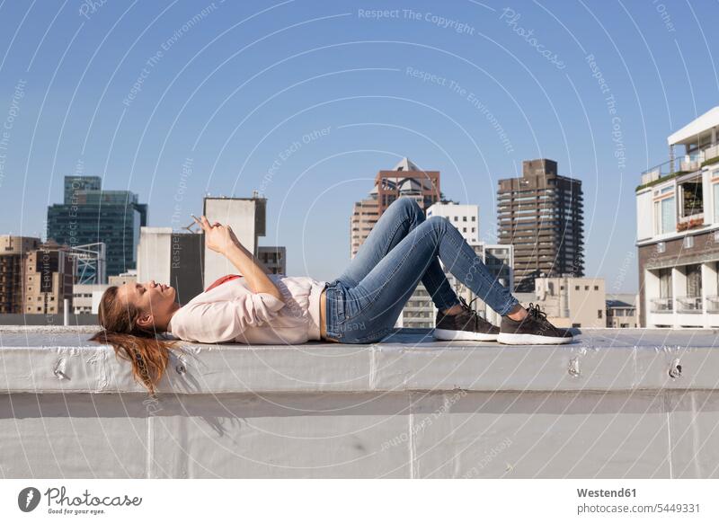 Young woman lying on balustrade of a rooftop terrace, using smart phone females women reading parapet blond blond hair blonde hair roof terrace deck laying down