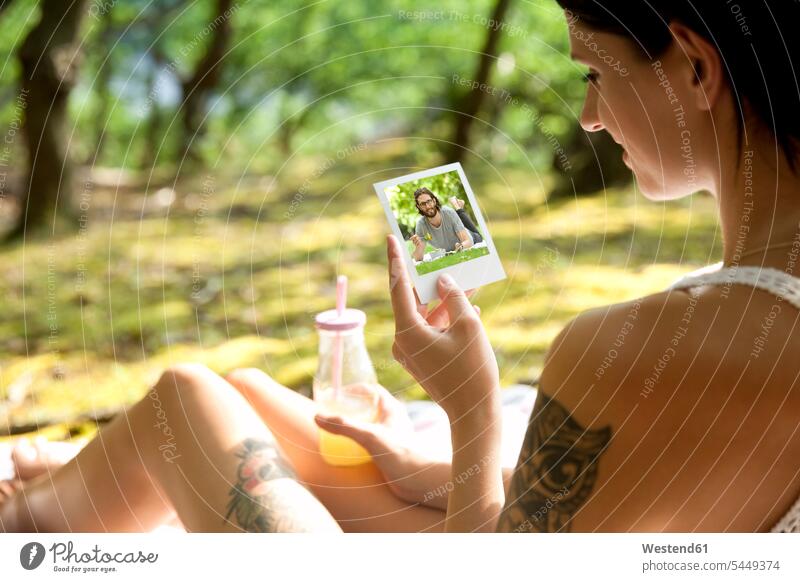 Young woman sitting on blanket in forest looking at instant photo Seated photograph photographs photos smiling smile females women eyeing image images picture