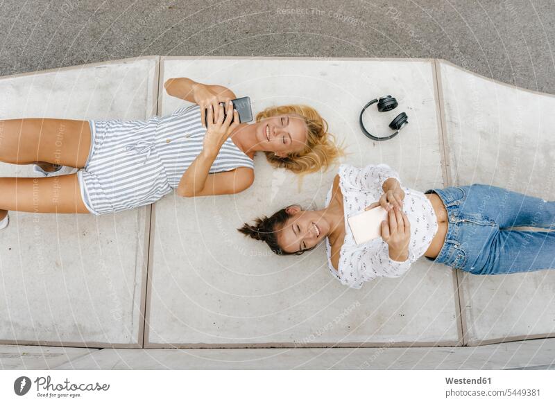 Two happy young women with cell phones lying on ramp in a skatepark female friends woman females mobile phone mobiles mobile phones Cellphone Ramp